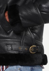 Chaqueta de cuero negra con puños y dobladillo de piel de oveja, que presenta herrajes dorados, detalles de hebilla y un acabado texturizado.