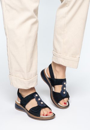 Feet in beige corduroy pants wearing black open-toe sandals with decorative pearls and adjustable straps, with red toenail polish visible.