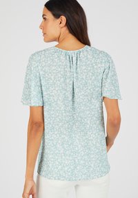Lichtblauwe blouse met korte mouwen en een wit bloemenpatroon, met een plooidetail bij de hals en een losse, vloeiende pasvorm.