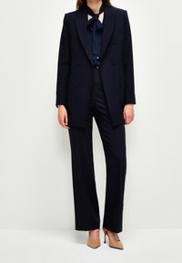 Navy double-breasted blazer over a dark blue blouse with a bow tie, paired with tailored navy trousers and light brown pointed heels.