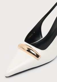 Chaussure slingback blanche à bout pointu avec intérieur noir et barre décorative dorée brillante à l'avant.