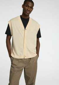 Bežna usnjena brezrokavnica z V-izrezom, kratkimi rokavi in gumbom, prekrita s črnim T-shirtom; kombinirana s svetlo rjavimi hlačami.