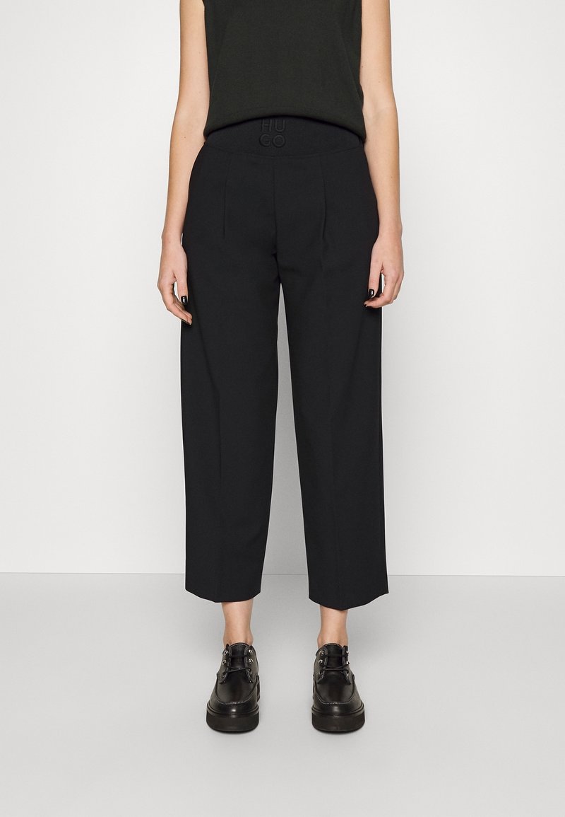 Personne portant un pantalon large noir raccourci, un haut noir sans manches et des chaussures noires à lacets, debout devant un fond blanc.