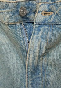 Vaaleansininen denimi, jossa on vetoketjukiinnitys ja kuparinvärinen nappi. Kankaassa on haalistunut pinta ja erottuva beige ommel.