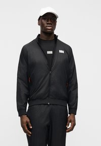 Μαύρο αδιάβροχο μπουφάν με μοτίβο σε πλέγμα και τσέπες με φερμουάρ. Διαθέτει ριμπ μανσέτες και κάτω μέρος, συνδυασμένο με μαύρο T-shirt.