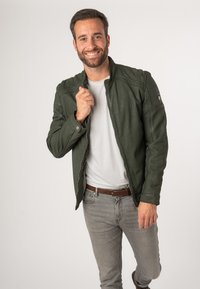 Groene, met rits gesloten jas met een gestructureerde afwerking, voorzien van gestikte accenten en een staande kraag, gedragen over een wit T-shirt en grijze jeans.