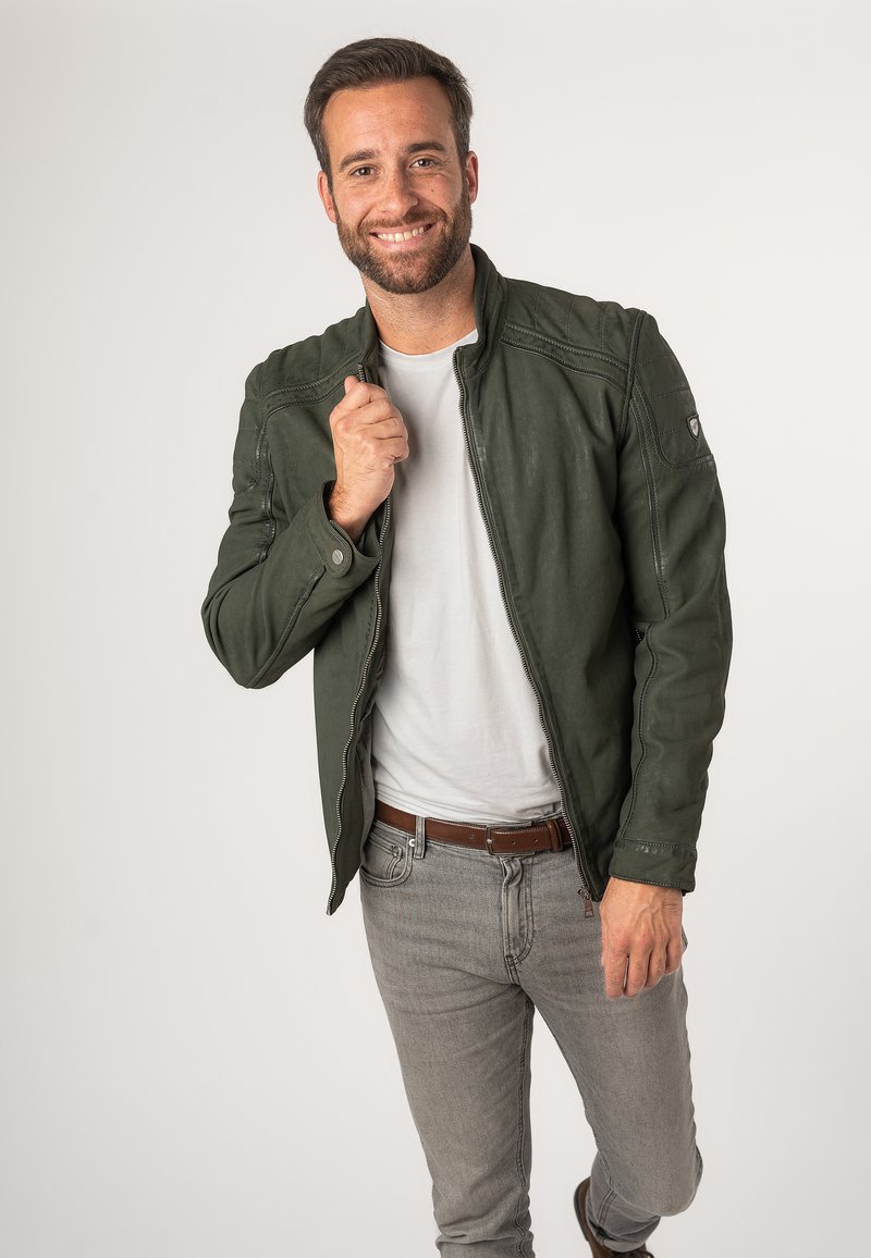 Groene, met rits gesloten jas met een gestructureerde afwerking, voorzien van gestikte accenten en een staande kraag, gedragen over een wit T-shirt en grijze jeans.