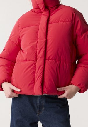 Person trägt eine leuchtend rote Steppjacke, hält den Saum mit den Händen und kombiniert dazu dunkelblaue Denim-Jeans vor einfarbigem Hintergrund.