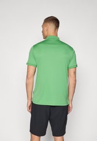 Grøn polo-shirt lavet af let stof med krave, korte ærmer og lige kant. Båret med sorte shorts.