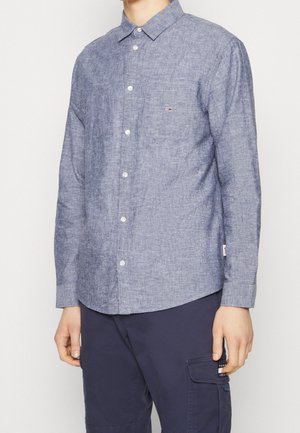 Hombre con camisa de manga larga azul claro abotonada, con bolsillo en el pecho y pantalones cargo azul marino oscuro.