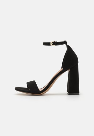 Sandale à talon haut en daim noir avec une plateforme, dotée d'une large bride à l'avant et d'une bride de cheville réglable, posée sur un talon bloc géométrique.