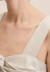 Haut en tissu beige clair avec de larges bretelles, présentant un détail noué au niveau de la poitrine. La texture semble douce et légèrement texturée.