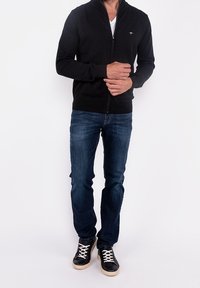 Černá zipová mikina, úzký střih, spárovaná s modrými džínami a černými teniskami s bílými tkaničkami. Jednoduchý design s malým logem na hrudi.