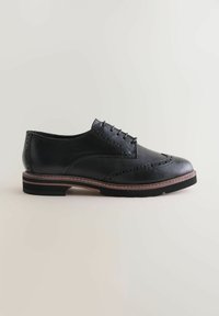 Čierne kožené brogues s dizajnom wingtip, hnedou vrstvenou podrážkou a čiernymi šnúrkami na svetlom neutrálom pozadí.