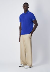 Blaues Poloshirt mit weißem Kragen, kombiniert mit beigen weit geschnittenen Hosen und weißen Sneakers. Einfaches, lässiges Outfit mit kontrastierenden Farben.