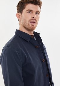 Giacca in cotone blu navy con chiusura a bottoni, colletto aperto e tasca sul petto. Caratterizzata da bottoni marroni e una superficie liscia.