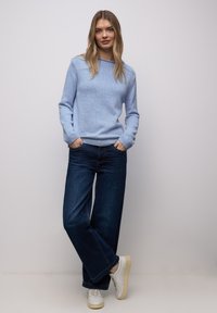 Femme aux longs cheveux blonds, debout contre un mur clair, portant un pull bleu clair, un jean large foncé et des baskets blanches.