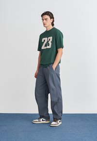 Grønn t-skjorte med korte ermer og lyst beige nummer "23" design, kombinert med grå avslappede bukser og svarte, hvite og grå sneakers.