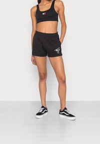 Černý sportovní bralet a černé teplákové šortky s detaily loga. Nohavice doplněné černobílými teniskami. Hladká tkanina, volný střih.