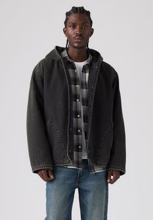 Giovane uomo con i capelli intrecciati che indossa una giacca nera con cappuccio sopra una camicia a quadri bianca e nera e jeans blu su uno sfondo semplice.