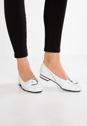 Białe skórzane baleriny z okrągłą srebrną klamrą, przeszyciami i niskim czarnym obcasem. Noszone z czarnymi dopasowanymi spodniami.