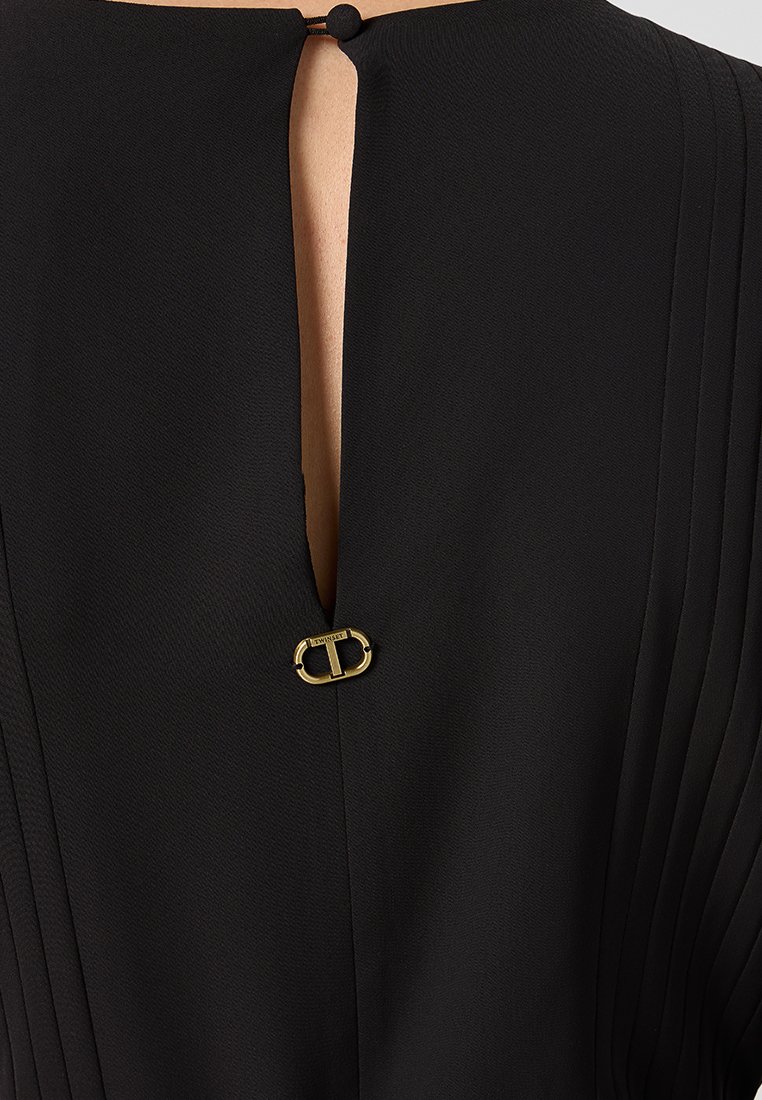 Abito nero con design a buchino sulla schiena, accentuato da una chiusura in metallo dorato e dettagli con pieghe lungo la schiena.