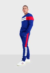 Tuta blu con accenti rossi, con una banda logo bianca sul petto e sui lati. Abbinata a sneakers bianche. Design slim fit.