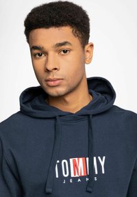 Sudadera azul marino con un logo frontal "TOMMY JEANS" en blanco y rojo, con cordones y tejido texturizado, sobre un fondo claro.