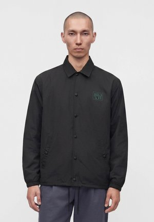 Homme à la tête rasée portant une veste noire boutonnée avec le logo "Maison Kitsuné" sur la poitrine et un pantalon gris, debout devant un fond uni.