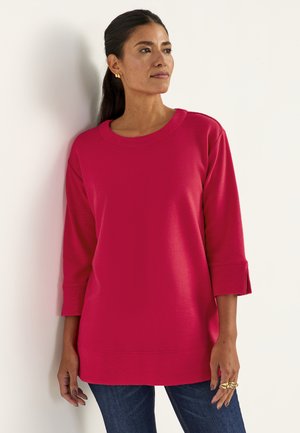 Fuchsiafarbener Langarmoberteil mit gerippter Textur, rundem Ausschnitt und Schlitzbündchen, kombiniert mit dunkelblauen Jeans. Keine sichtbaren Logos oder Muster.