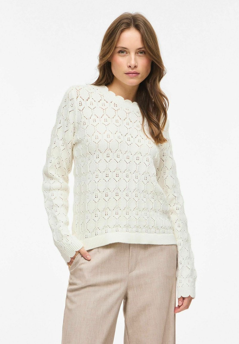 Femme portant un pull en maille blanc à motifs avec des bords festonnés et un pantalon beige, debout, une main dans la poche.