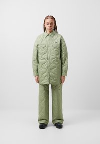 Jachetă verde matlasată cu închidere cu nasturi și două buzunare pe piept, asortată cu pantaloni matlasati. Prezentă un detaliu discret cu logo.