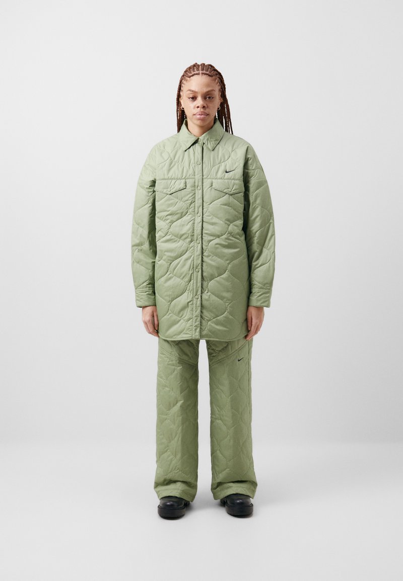 Jachetă verde matlasată cu închidere cu nasturi și două buzunare pe piept, asortată cu pantaloni matlasati. Prezentă un detaliu discret cu logo.