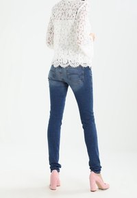 Top long en dentelle blanche à manches longues avec un ourlet festonné, associé à un jean bleu ajusté et des chaussures à talons hauts roses pâles, vu de dos.