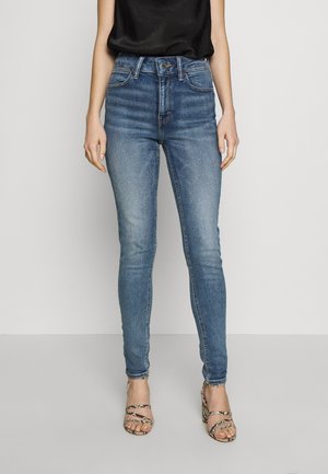 Vrouw draagt hoge, slim-fit blauwe jeans met vervaagde details, een zwart mouwloos topje en sandalen met hak met slangenhuidpatroon.