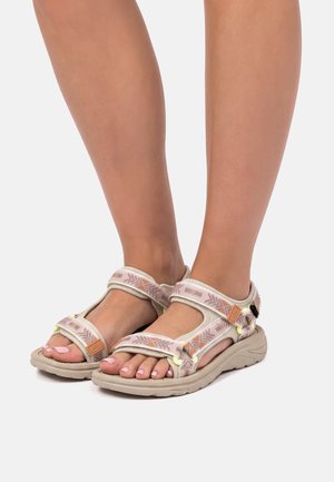 Füße in beige-rosa gemusterten Outdoor-Sandalen mit verstellbaren Riemen vor hellem Hintergrund.