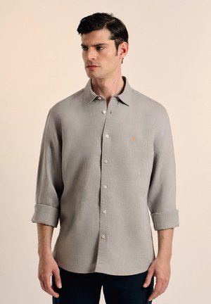 Camisa de botón de color gris claro en una tela texturizada, con cuello clásico, puños enrollados y un pequeño emblema bordado en el pecho.