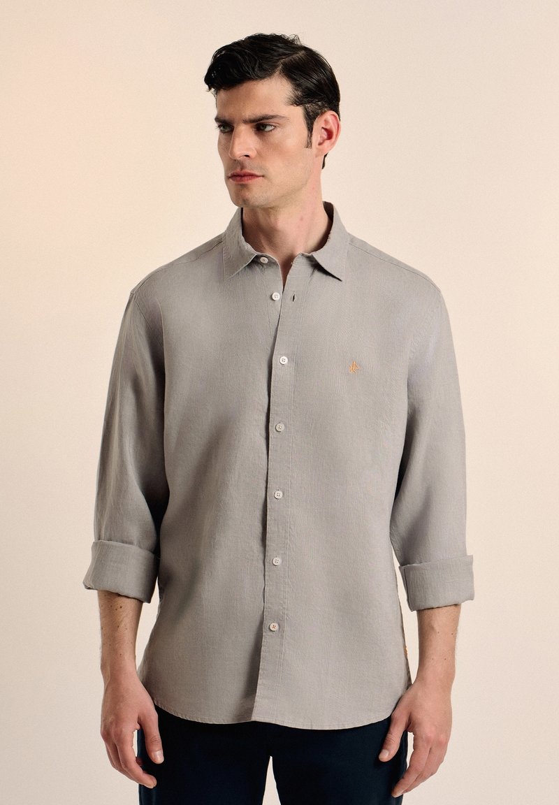 Camisa de botón de color gris claro en una tela texturizada, con cuello clásico, puños enrollados y un pequeño emblema bordado en el pecho.