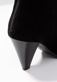 Czarne zamszowe botki za kostkę z wysmukłym, wysokim obcasem. Posiadają boczny zamek błyskawiczny i gładką fakturę, podkreślając elegancki i nowoczesny design.