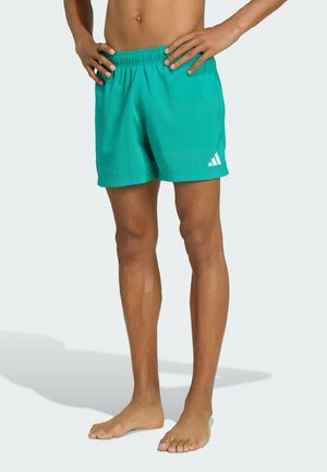 Osoba boso stojąca z rękami na biodrach, ubrana w jaskrawo turkusowe spodenki Adidas z białym logo na jasnym tle.