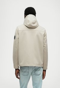 Bežna jakna s kapuco z gladko teksturo, ki ima oznako na levem rokavu in preprost, elegantno oblikovan kroj. Nosijo jo nad svetlo modrimi kavbojkami.