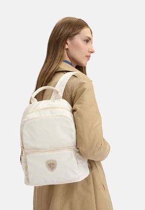 Donna con capelli lunghi in cappotto beige che indossa uno zaino color crema con tasca frontale e cerniere dorate, rivolta verso destra su sfondo chiaro.