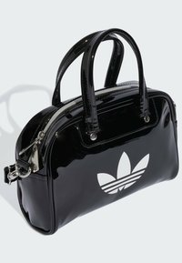 Sac à main noir en vernis avec double poignées, fermeture éclair argentée et un grand logo Adidas trefoil blanc sur le devant. Forme rectangulaire. Texture brillante.