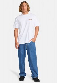 T-shirt branca de algodão com mangas curtas e um pequeno logótipo circular no peito, combinada com jeans denim azuis de corte largo.