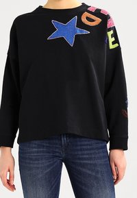 Czarny sweatshirt z długimi rękawami, zdobiony kolorowymi aplikacjami w kształcie gwiazdek i liter w odcieniach niebieskiego, różowego, zielonego i pomarańczowego po jednej stronie.