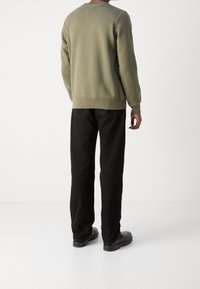 Uomo che indossa un felpa verde oliva, jeans neri a gamba dritta e scarpe in pelle nere, in piedi davanti a uno sfondo bianco semplice.