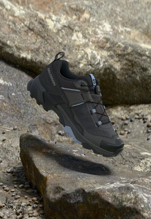 Zwarte Salomon trailrunningschoen met robuuste zool, hangend boven rotsachtig terrein met grote stenen in een natuurlijke buitenomgeving.