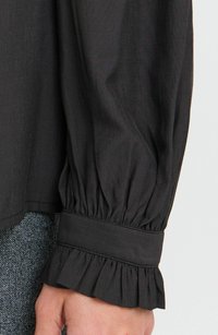 Schwarzes Langarmshirt mit gepufften, gerafften Ärmeln und einem Rüschensaum. Hergestellt aus strukturiertem Stoff mit dezenten horizontalen Streifen.