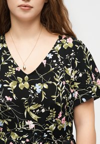 Γυναίκα που φοράει μαύρο φόρεμα με λουλούδια και φύλλα σε ροζ, μπλε και πράσινο χρώμα, και ένα χρυσό κολιέ με μενταγιόν σε σχήμα σταγόνας.