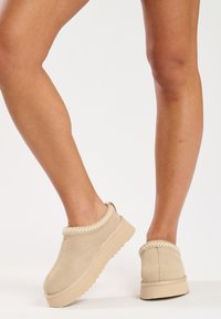 Beige slip-on shoes made of soft suede with a textured collar and a thick rubber sole. Features a rounded toe and minimal design details.